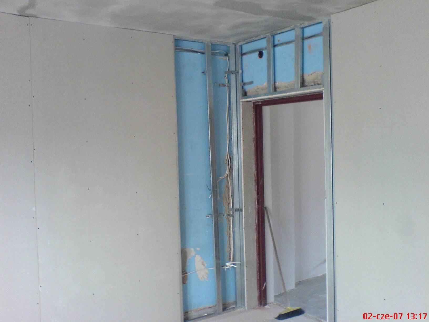 Montaż ścianki działowej: metalowy stelaż z niebieską izolacją, częściowo pokryty płytami gipsowo-kartonowymi, widoczne przewody elektryczne i szczotka na podłodze.