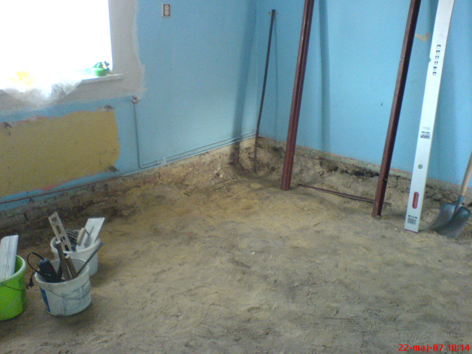 Przygotowanie podłoża pod wylewkę w rogu pomieszczenia z błękitnymi ścianami, widoczne narzędzia w wiadrach, poziomica oparta o ścianę, ślady po usuniętej starej podłodze.