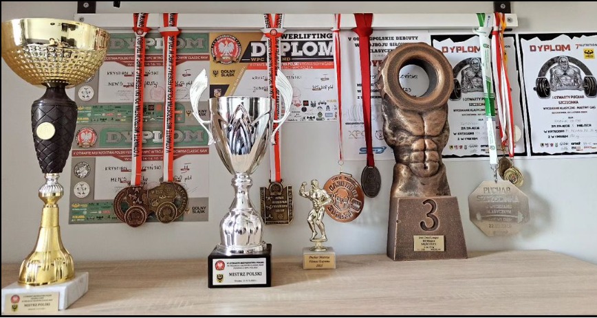 Półka z trofeami i medalami zdobytymi w zawodach siłowych, w tle dyplomy i plakaty z zawodów, widoczne puchary w kolorach złota i srebra, oraz statuetka kulturysty