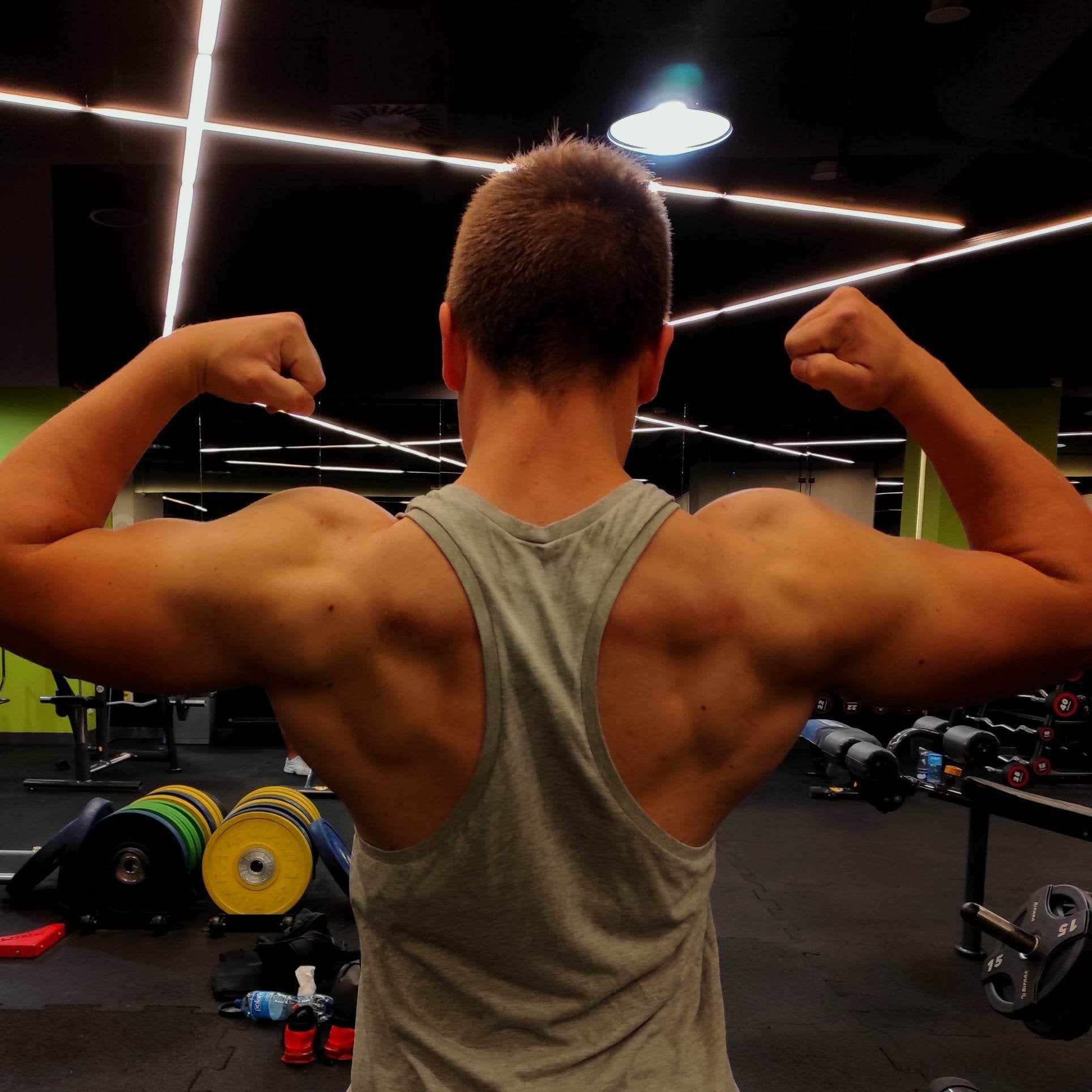Mężczyzna w szarej koszulce typu bokserka, z widocznymi mięśniami pleców i bicepsami, pozujący na tle siłowni z obciążeniami i maszynami.