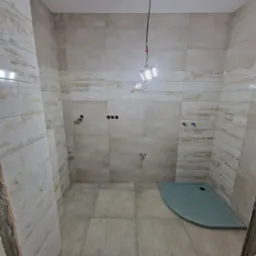 Łazienka w trakcie wykańczania, widoczne płytki imitujące drewno na ścianach, szary brodzik i tymczasowe oświetlenie budowlane.