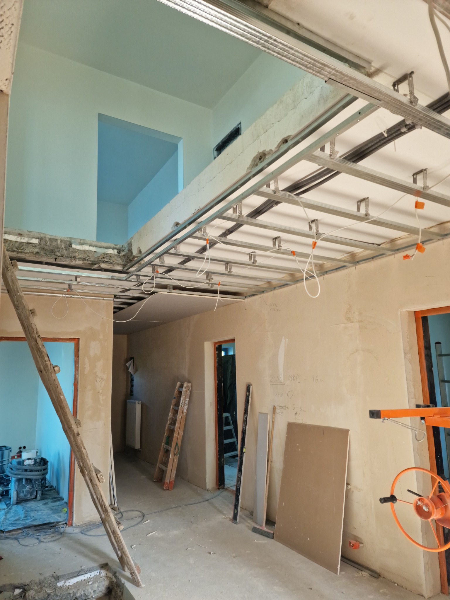 Wnętrze w trakcie montażu sufitu podwieszanego z widoczną konstrukcją metalową, przewodami elektrycznymi, częściowo otynkowanymi ścianami, schodami drewnianymi opartymi o framugę drzwi...