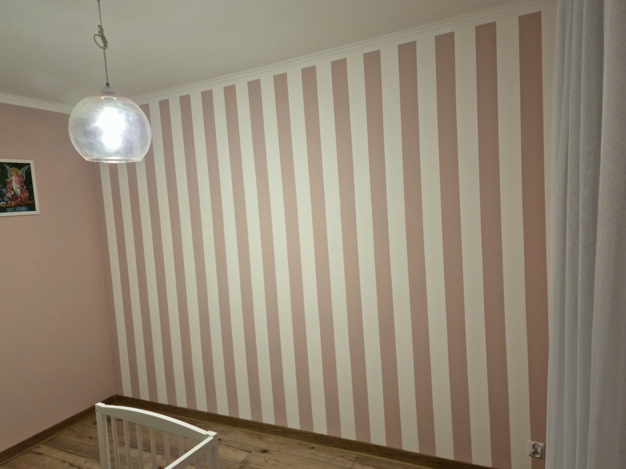 Ściana w pokoju dziecięcym pomalowana w pionowe pasy w odcieniach różu i bieli, widoczny fragment białego łóżeczka i lampa wisząca
