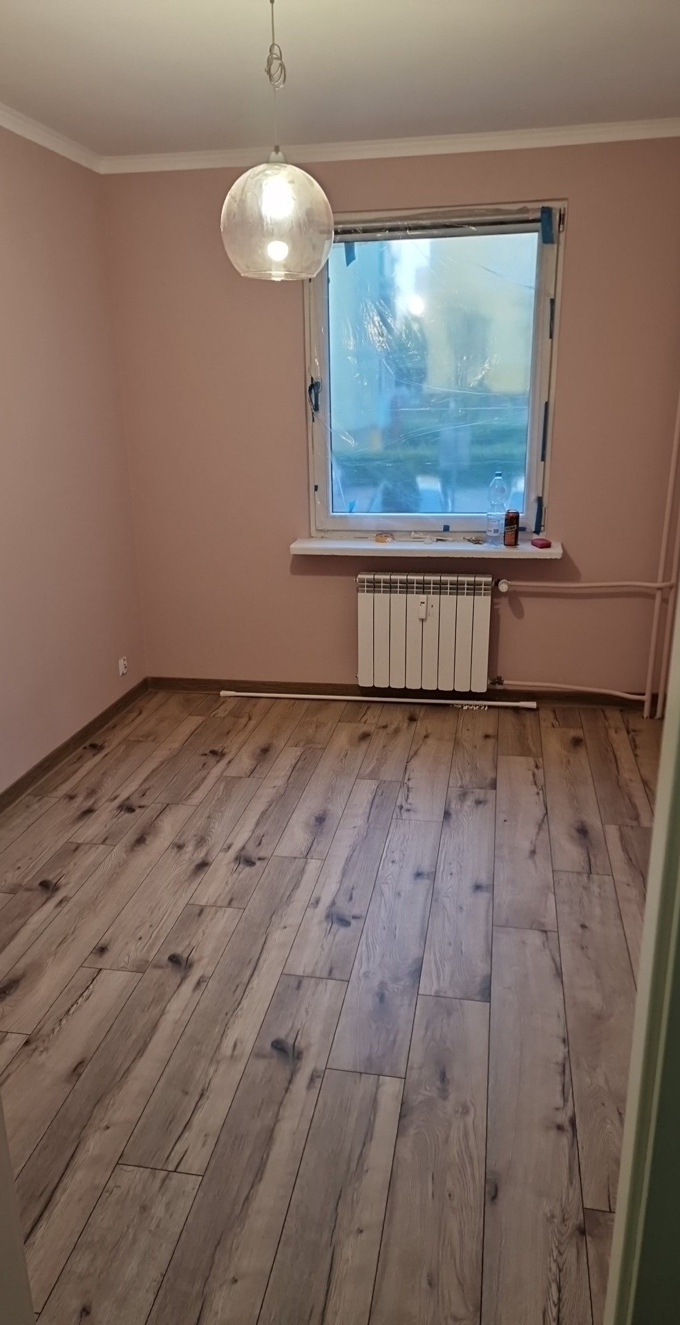 Pokój po odświeżeniu z drewnianą podłogą, ściany pomalowane na różowo, okno zabezpieczone folią malarską, biały grzejnik pod oknem, lampa wisząca w kształcie kuli.