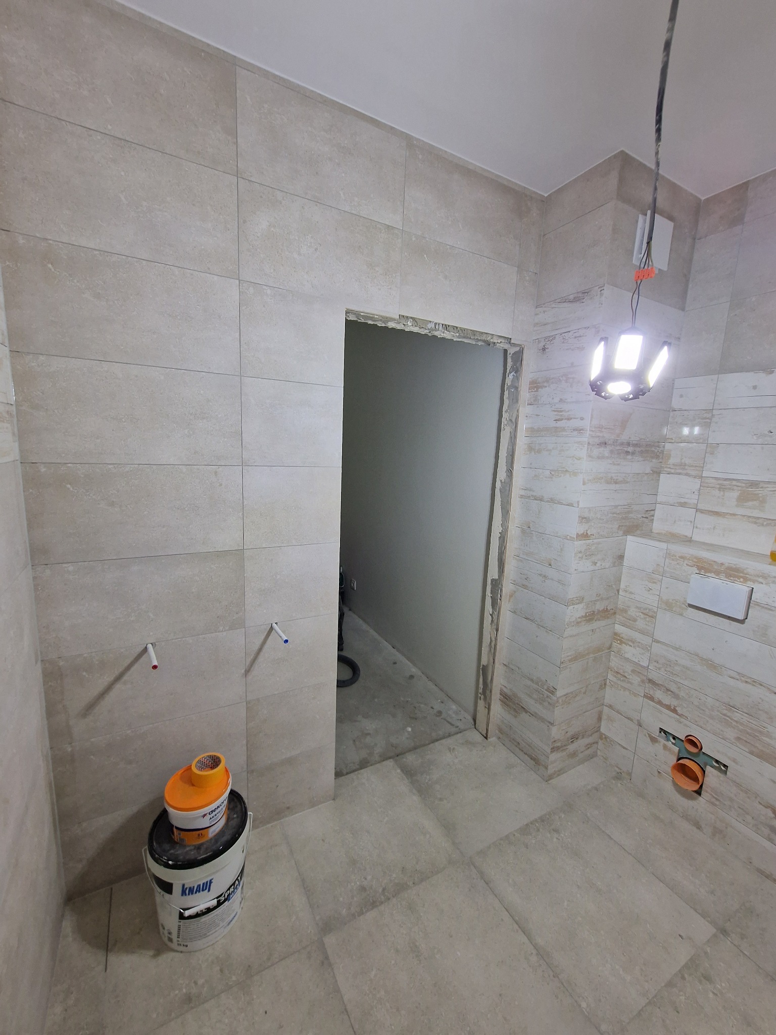 Łazienka w trakcie remontu: beżowe płytki ścienne i podłogowe, widoczne przyłącza wodne, wiadro z klejem Knauf oraz lampa budowlana.