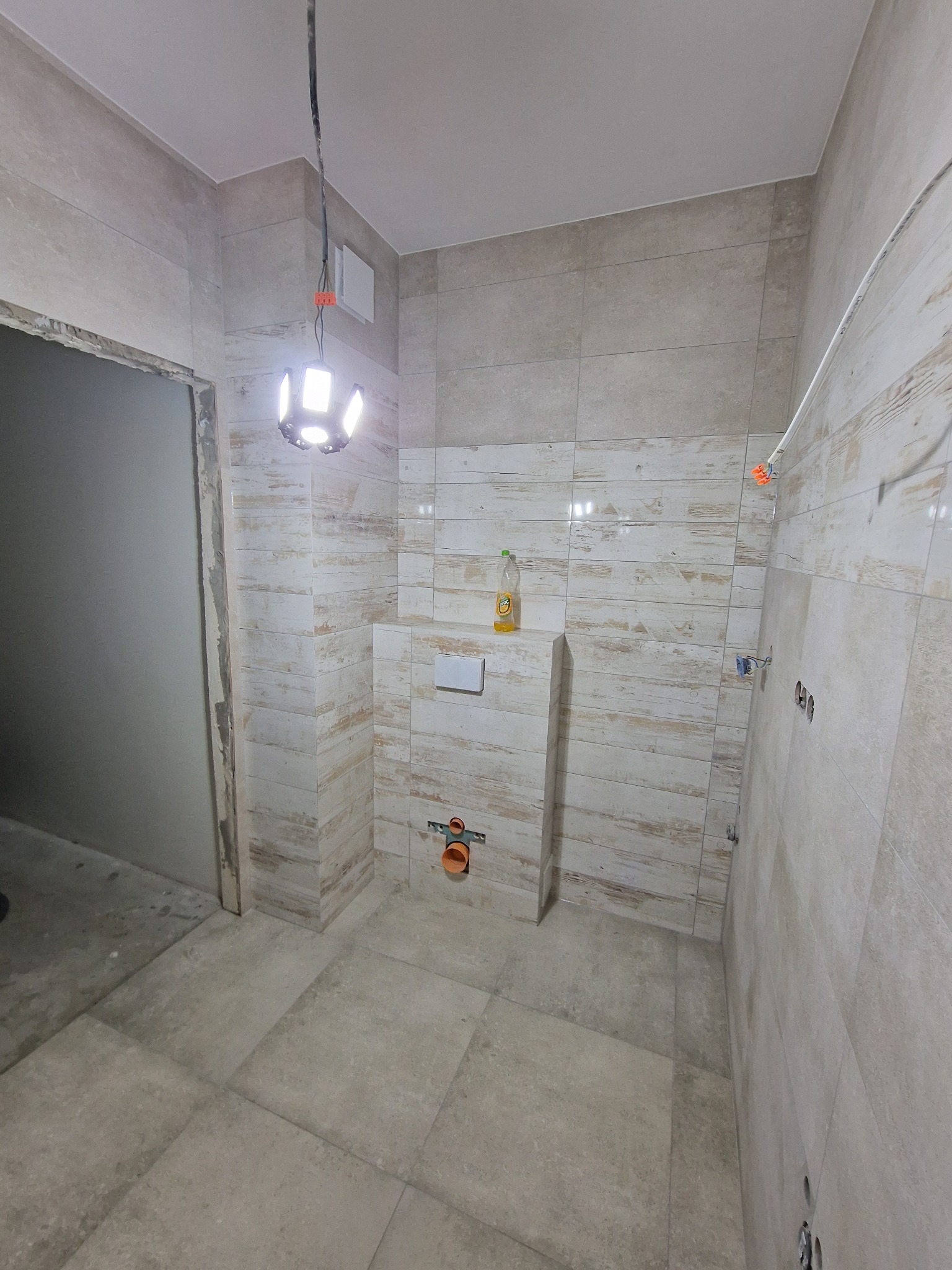 Wnętrze łazienki w trakcie prac wykończeniowych: ułożone beżowe płytki imitujące drewno na ścianach i podłodze, widoczna instalacja podtynkowego WC, butelka płynu na obudowie stelaża, wisząca lampa...