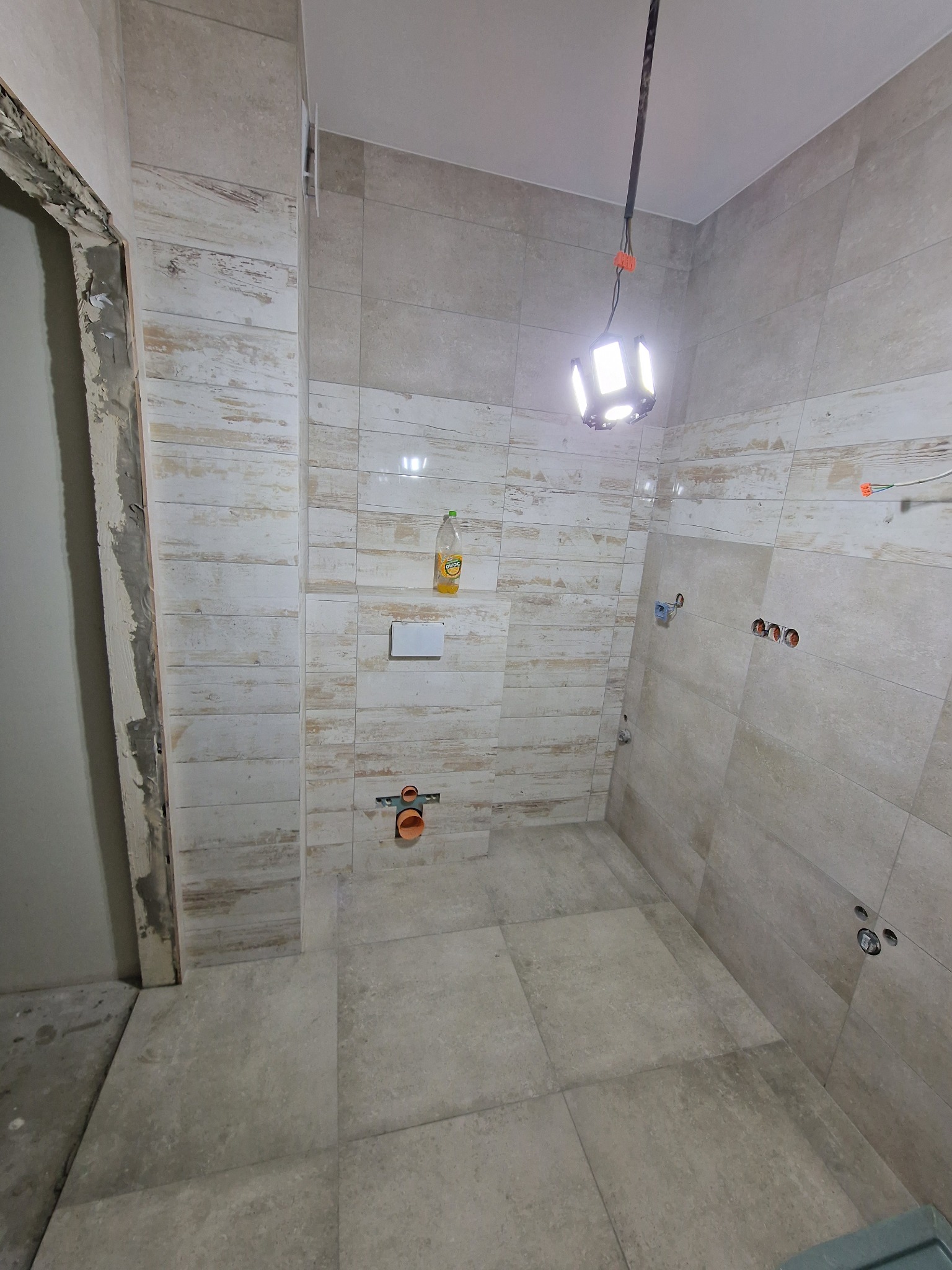 Wnętrze łazienki w trakcie prac wykończeniowych: ułożone płytki imitujące drewno na ścianach, szare płytki podłogowe, widoczne instalacje wodno-kanalizacyjne i elektryczne, oświetlenie robocze.