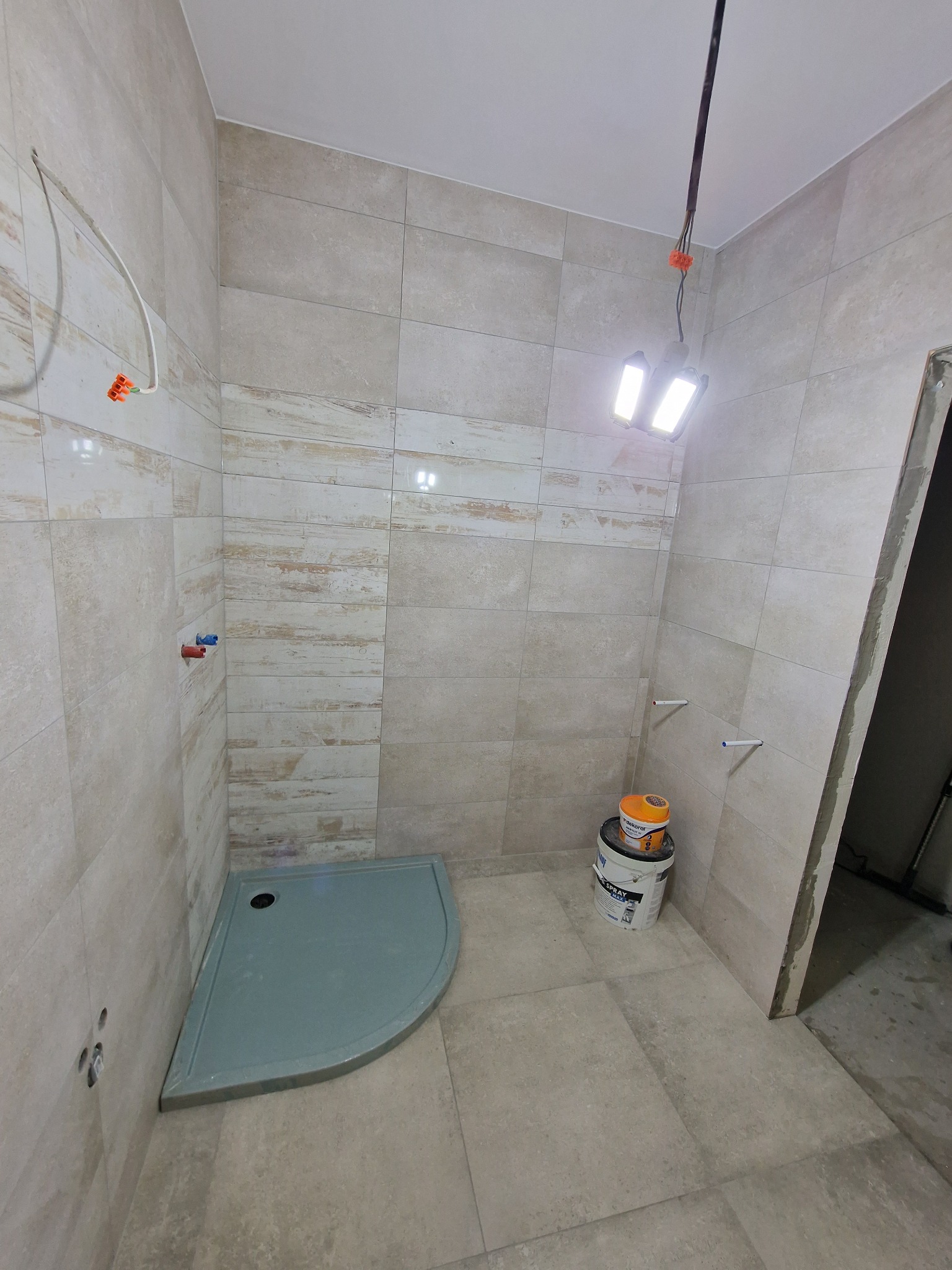 Wnętrze łazienki w trakcie remontu: ułożone beżowe płytki imitujące drewno, zamontowany szary brodzik, widoczne puszki z farbą i tymczasowe oświetlenie budowlane.