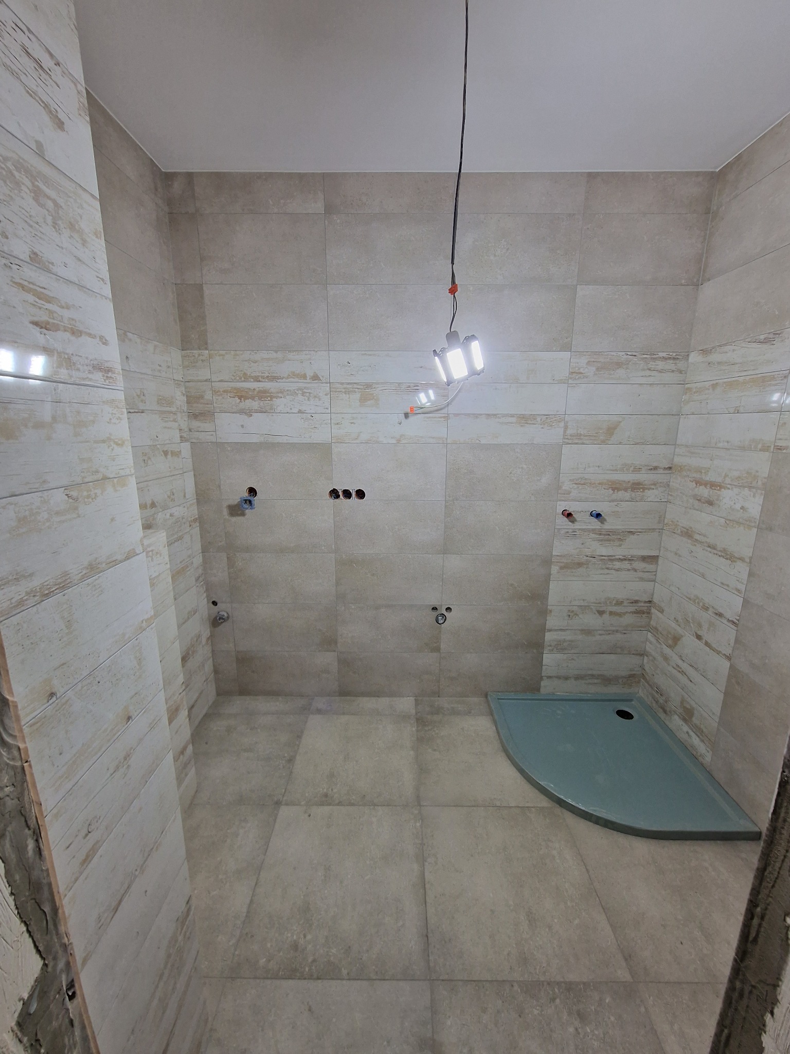 Łazienka w trakcie wykańczania, widoczne płytki imitujące drewno na ścianach, szary brodzik i tymczasowe oświetlenie budowlane.