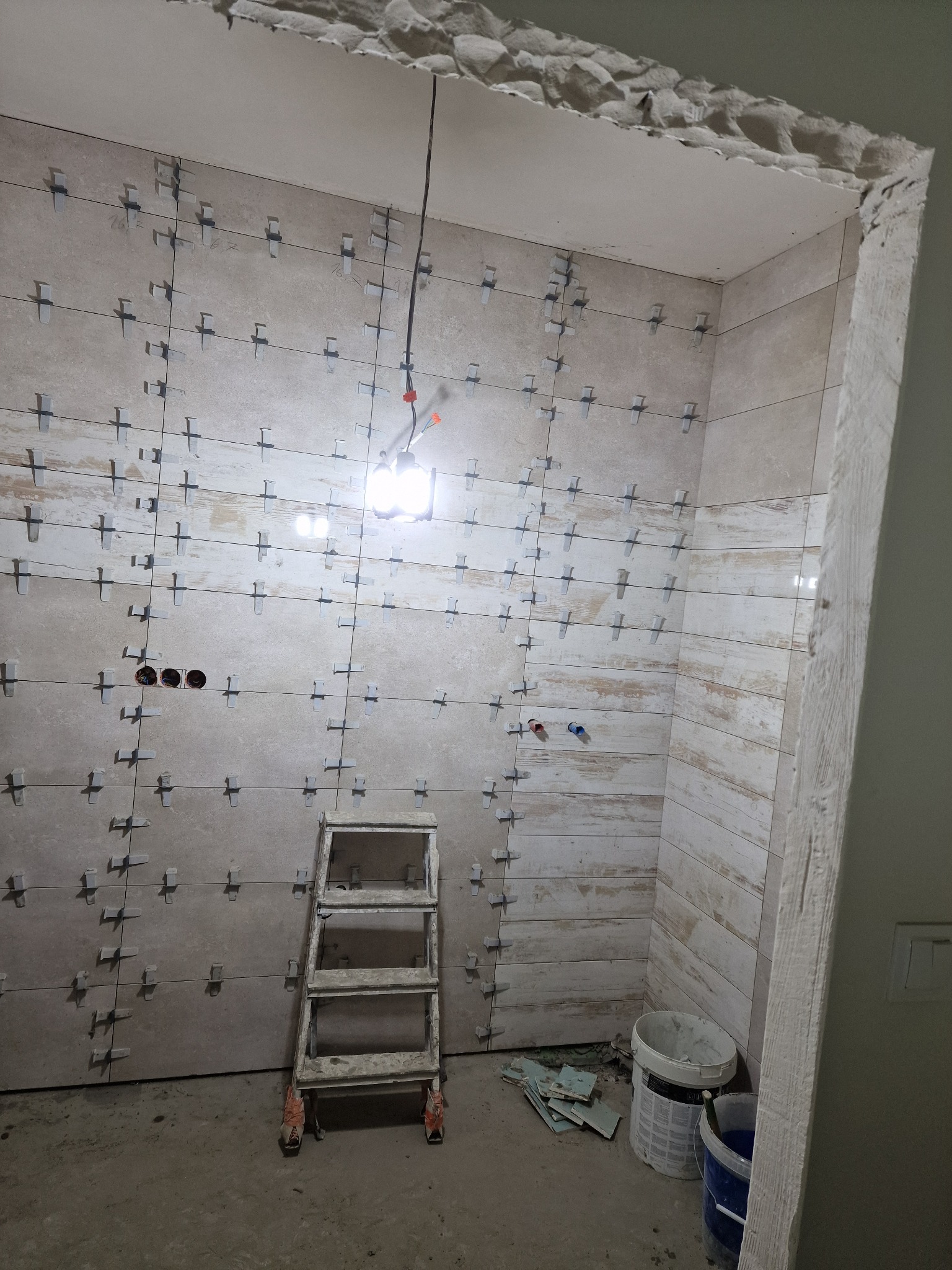 Łazienka w trakcie remontu: ułożone płytki z krzyżykami dystansowymi, drabina malarska, widoczne instalacje elektryczne i hydrauliczne, surowy stan wykończenia.