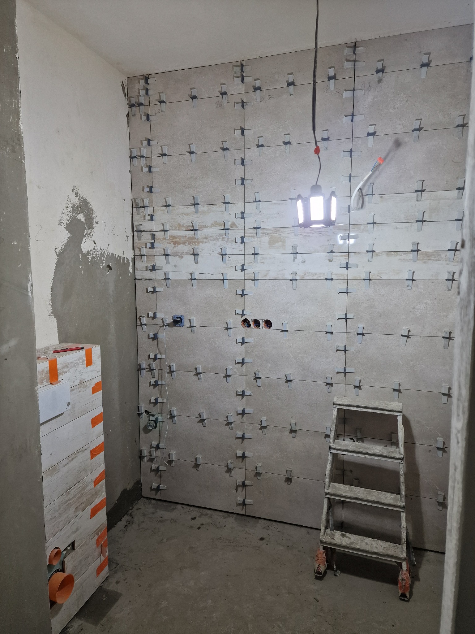 Ściana w trakcie układania płytek ceramicznych, widoczne krzyżyki dystansowe, drabina i tymczasowe oświetlenie budowlane.
