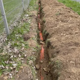Wykopany rów z pomarańczowymi rurami instalacyjnymi, widoczny wzdłuż ogrodzenia i drogi.