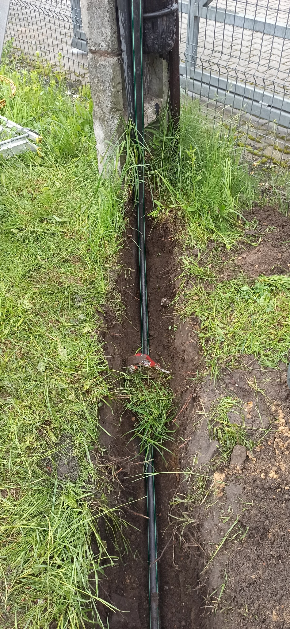 Czarna rura z zielonym paskiem ułożona w wykopie w ziemi porośniętej trawą, obok leży czerwono-srebrny nóż do cięcia rur, widoczny betonowy słupek ogrodzenia i metalowa siatka.