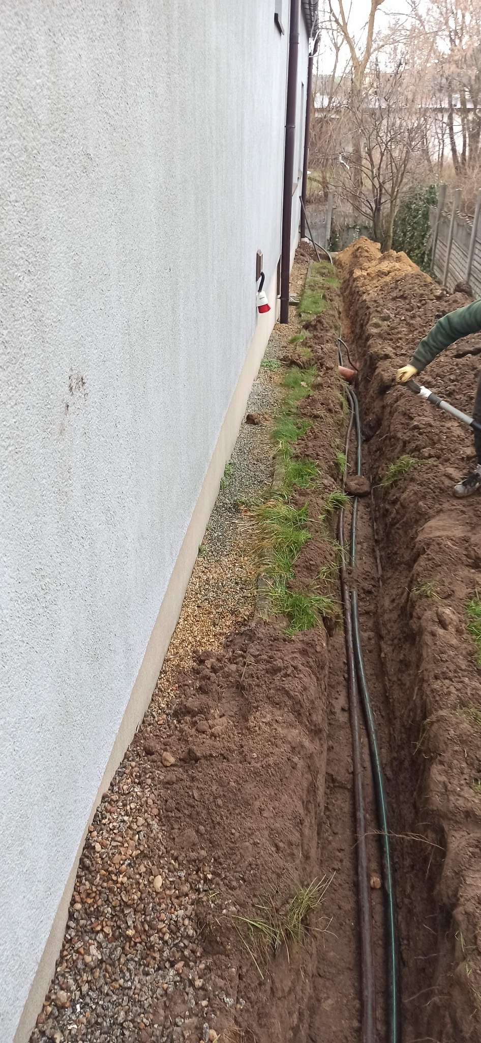 Wykopany rów wzdłuż ściany budynku z widocznymi kablami instalacyjnymi w ziemi, osoba w rękawicach roboczych używa szpadla do dalszych prac.