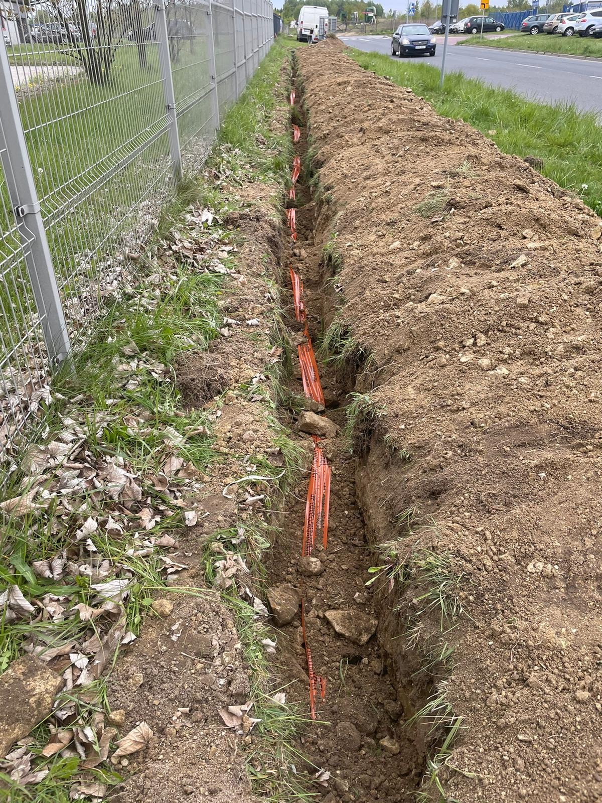 Wykopany rów z pomarańczowymi rurami instalacyjnymi, widoczny wzdłuż ogrodzenia i drogi.
