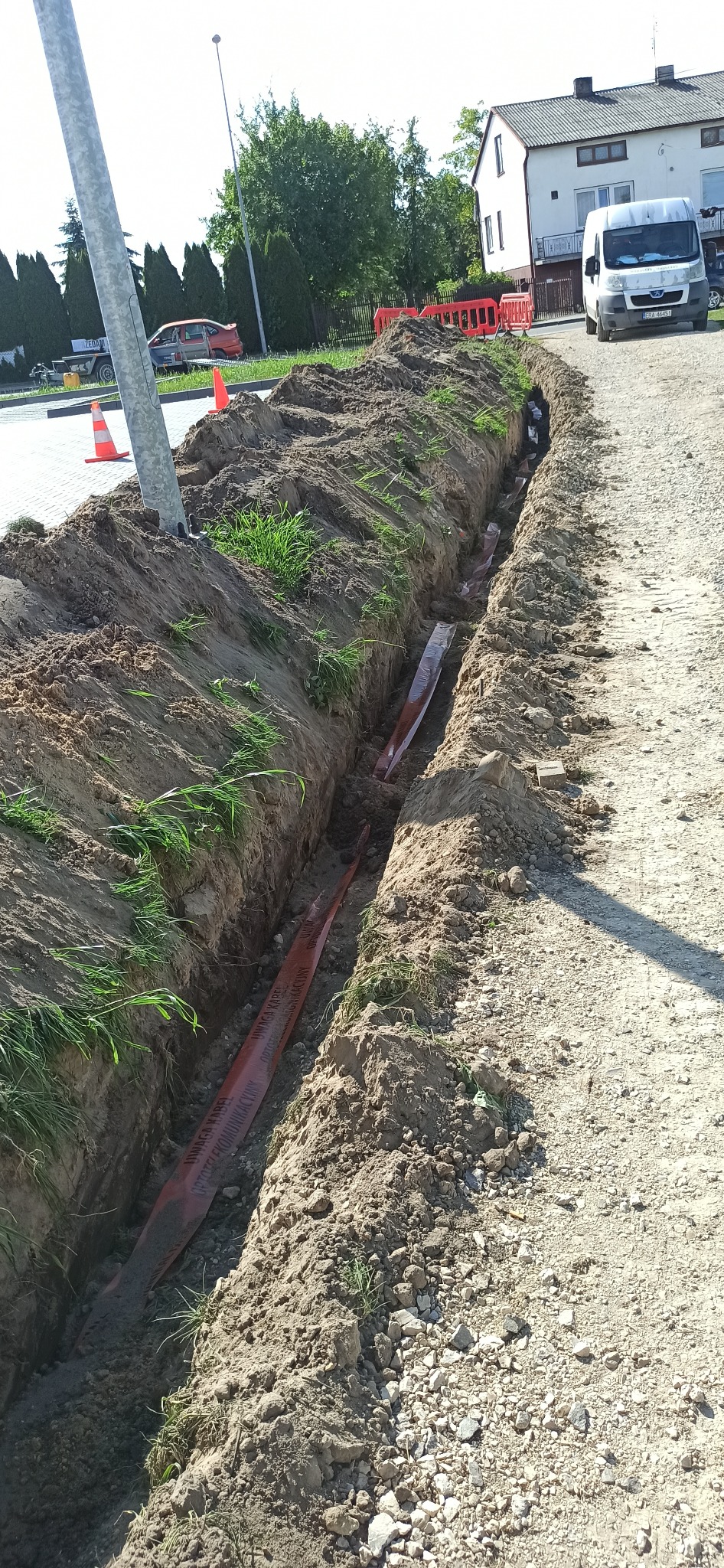 Wykopany rów w ziemi z pomarańczowym kablem telekomunikacyjnym w środku, widoczne trawy na krawędziach wykopu, słup oświetleniowy i zaparkowany biały van w tle.
