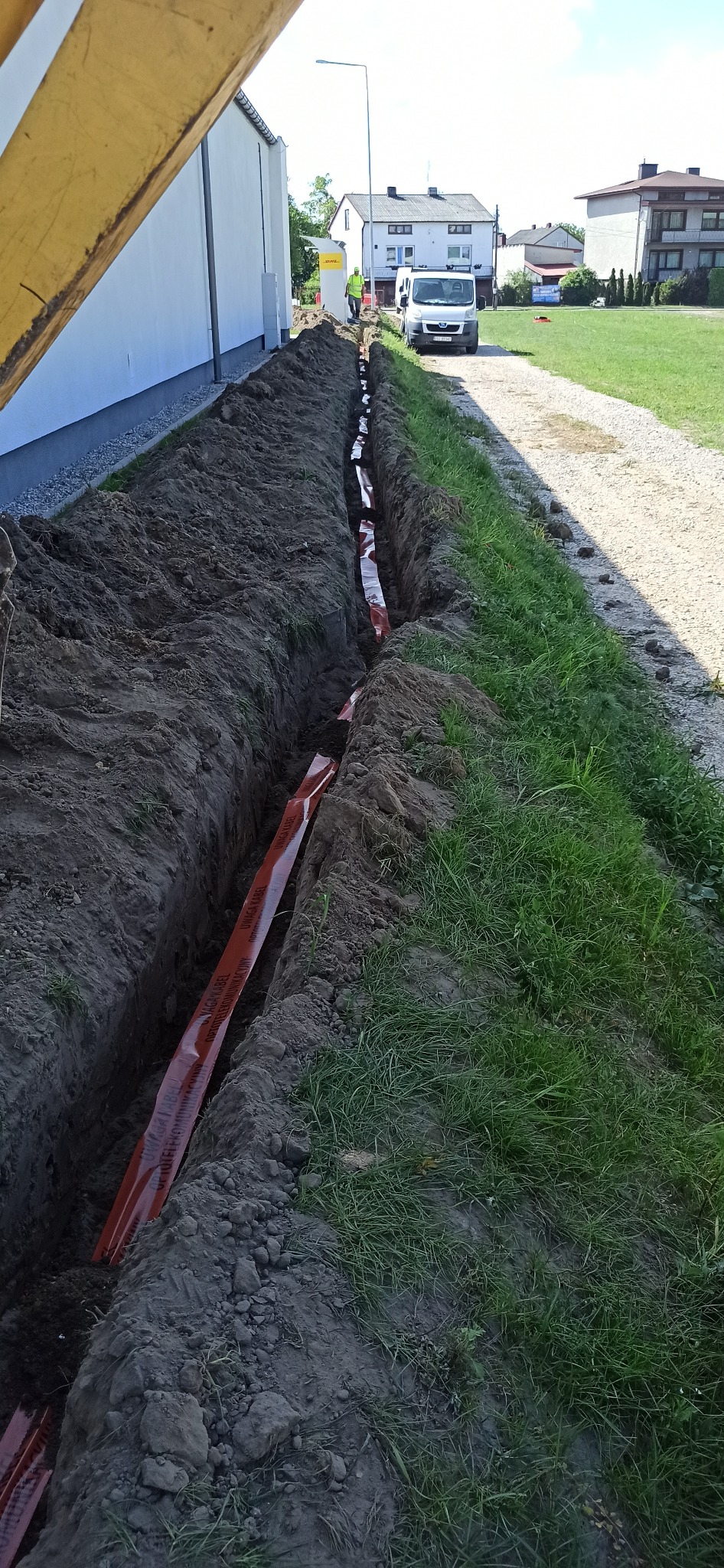 Wykopany rów z pomarańczową taśmą ostrzegawczą 'Uwaga Kabel Optyczny Telekomunikacyjny' ułożoną wzdłuż jego dna, widoczny pracownik w odblaskowej kamizelce i biały samochód dostawczy w tle.