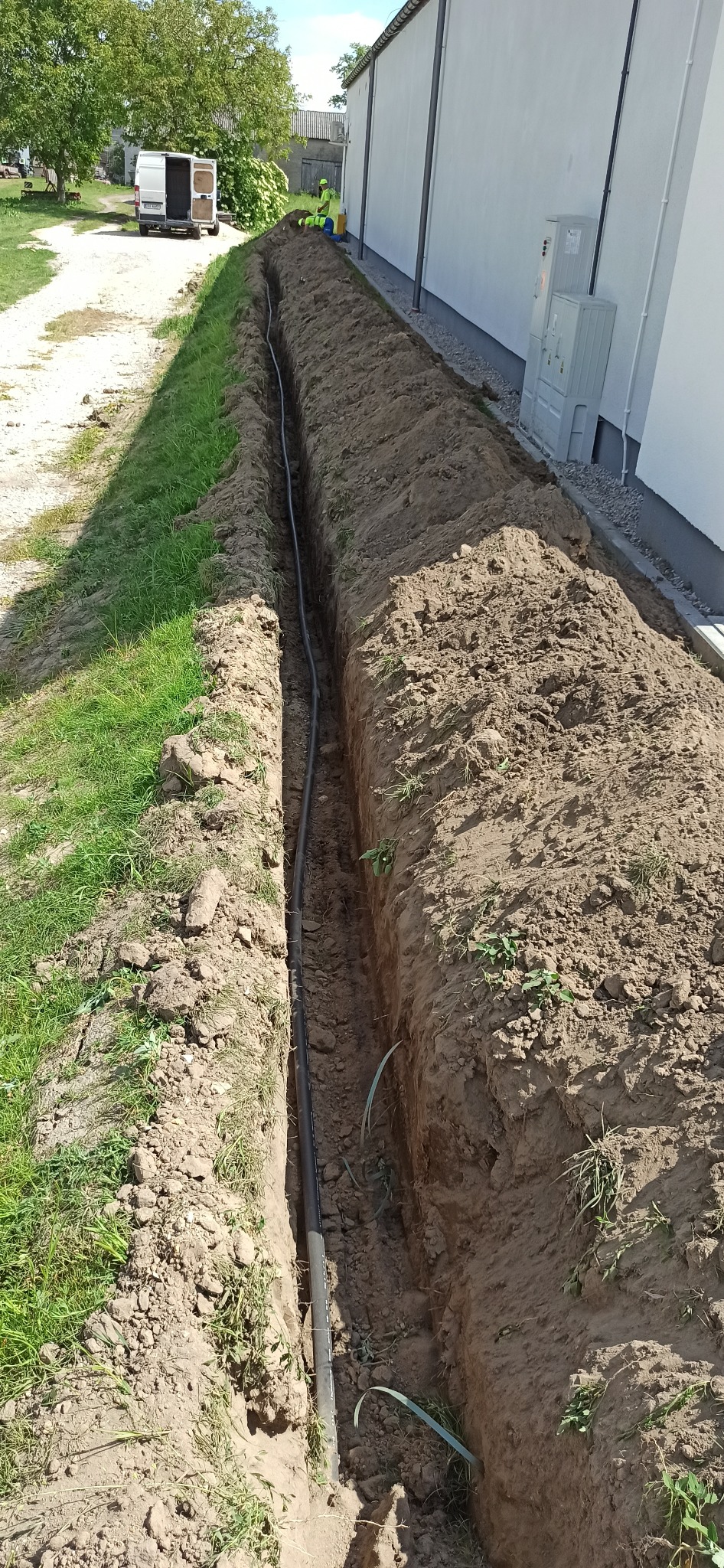 Wykopany rów z ułożonym kablem wzdłuż ściany budynku, widoczny pracownik w odblaskowej kamizelce i bus na tle drzew.