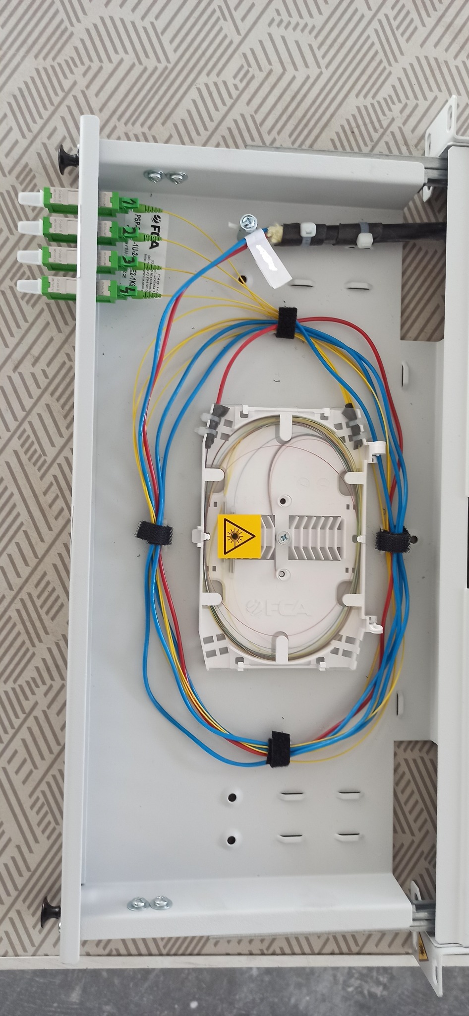 Otwarta skrzynka telekomunikacyjna z uporządkowanymi kablami światłowodowymi w kolorach niebieskim, czerwonym i żółtym, z widocznym ostrzeżeniem o promieniowaniu laserowym.