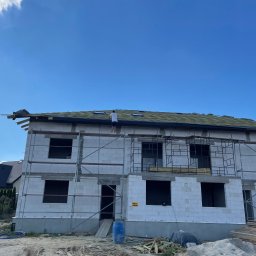 NKF HOUSE SPÓŁKA Z OGRANICZONĄ ODPOWIEDZIALNOŚCIĄ - Wznoszony budynek z białego bloczka na etapie prac dekarskich, widoczne rusztowania i dekarz na dachu, błękitne niebo w tle.