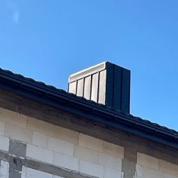 NKF HOUSE SPÓŁKA Z OGRANICZONĄ ODPOWIEDZIALNOŚCIĄ - Niski kąt ukazuje czarny, metalowy komin na dachu budynku z białego bloczka, na tle błękitnego nieba.