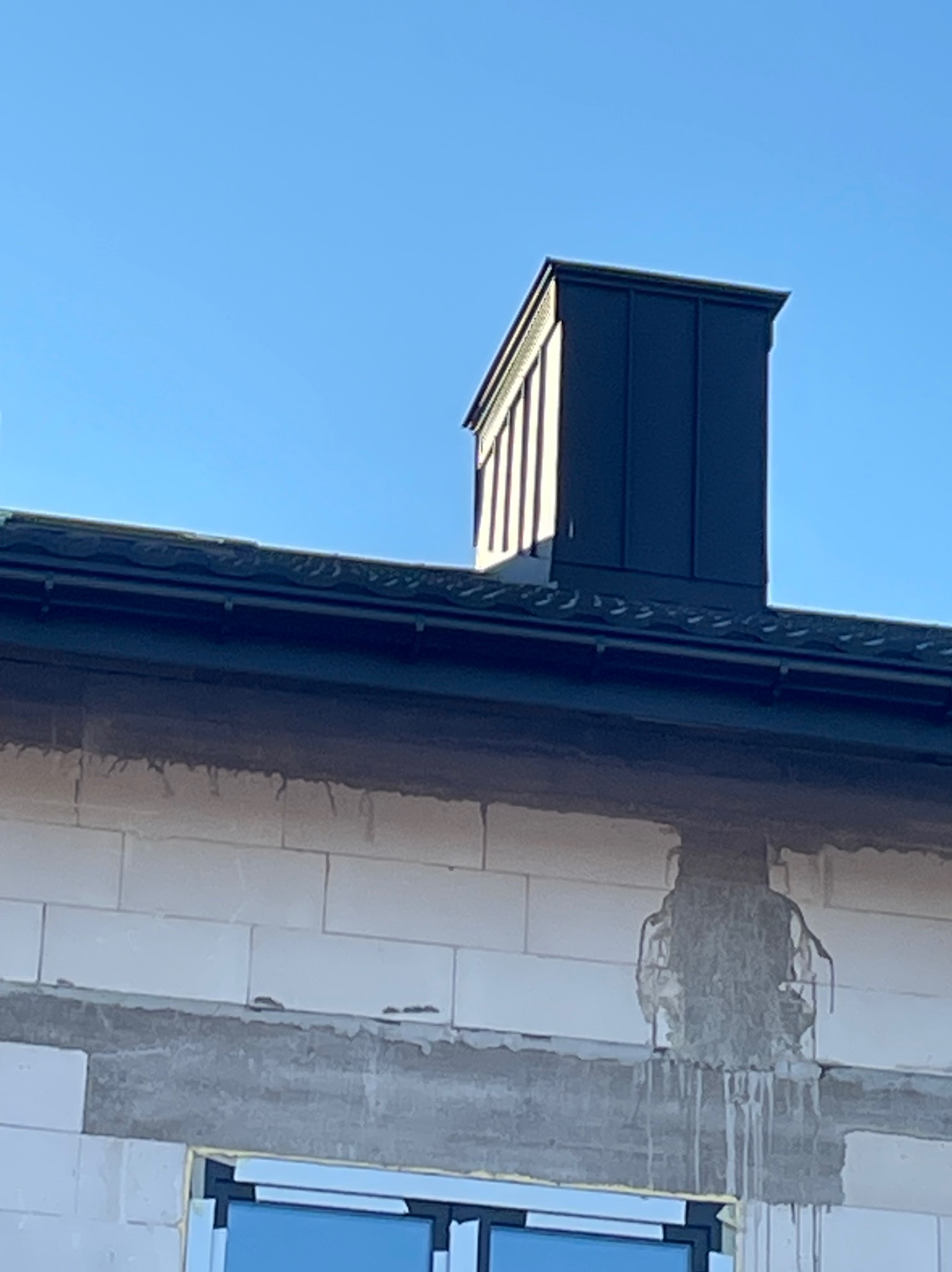 Szary komin z blachy na tle błękitnego nieba, widoczny fragment dachu z rynnami i mur z betonu komórkowego w trakcie budowy.