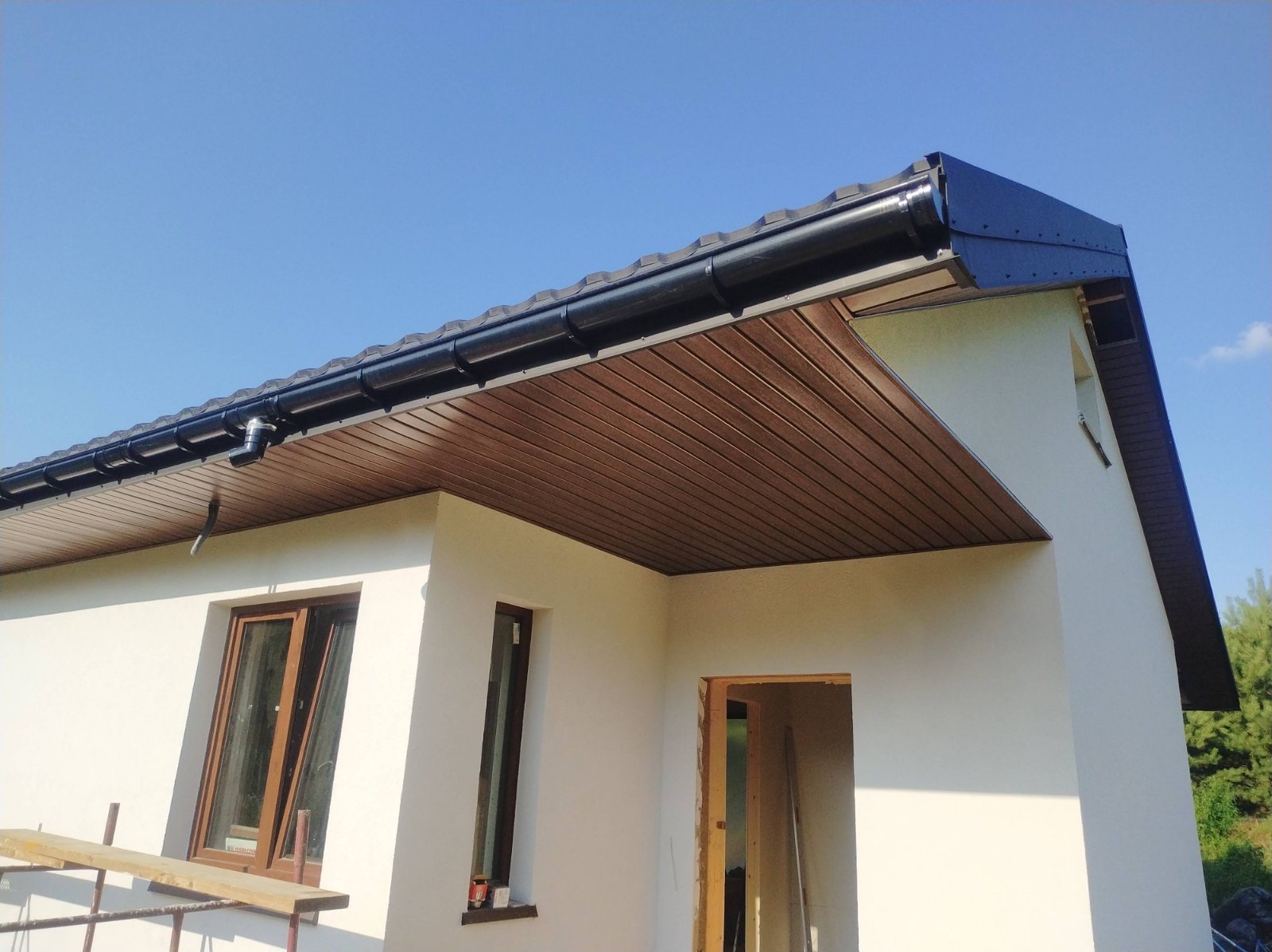 Fragment nowo wybudowanego domu z zamontowanymi, czarnymi rynnami i podbitką w kolorze brązowym, widoczne okna i niezakończony otwór drzwiowy.