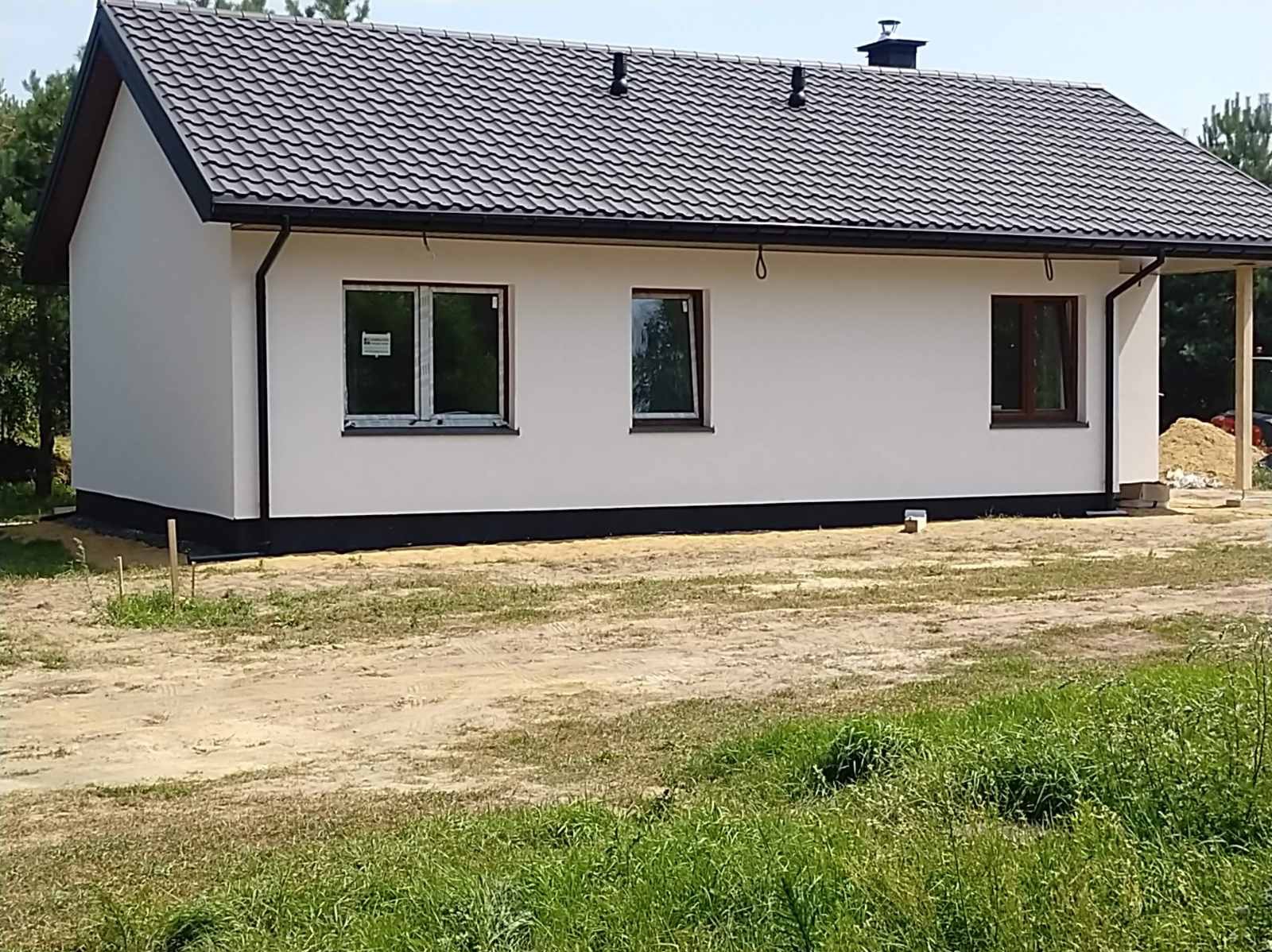 Gotowy, parterowy dom z jasną elewacją, ciemną podmurówką i dachem z blachodachówki w kolorze grafitowym, widoczne okna z brązowymi ramami i fragment zadaszonego tarasu w surowym stanie.