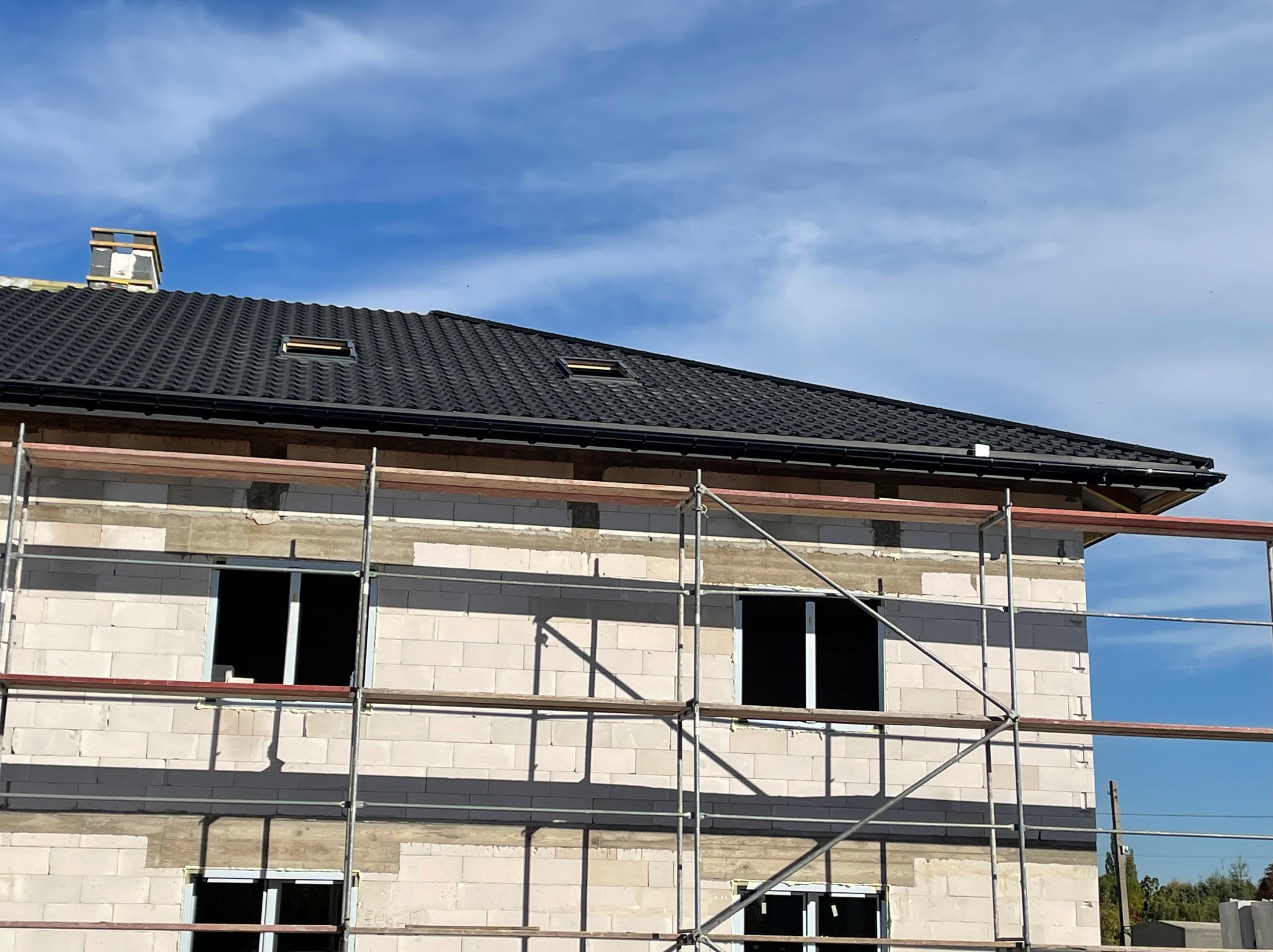 Budowa domu z białych bloczków, z zamontowaną czarną dachówką i rynnami, widoczne rusztowania i okna bez szyb.