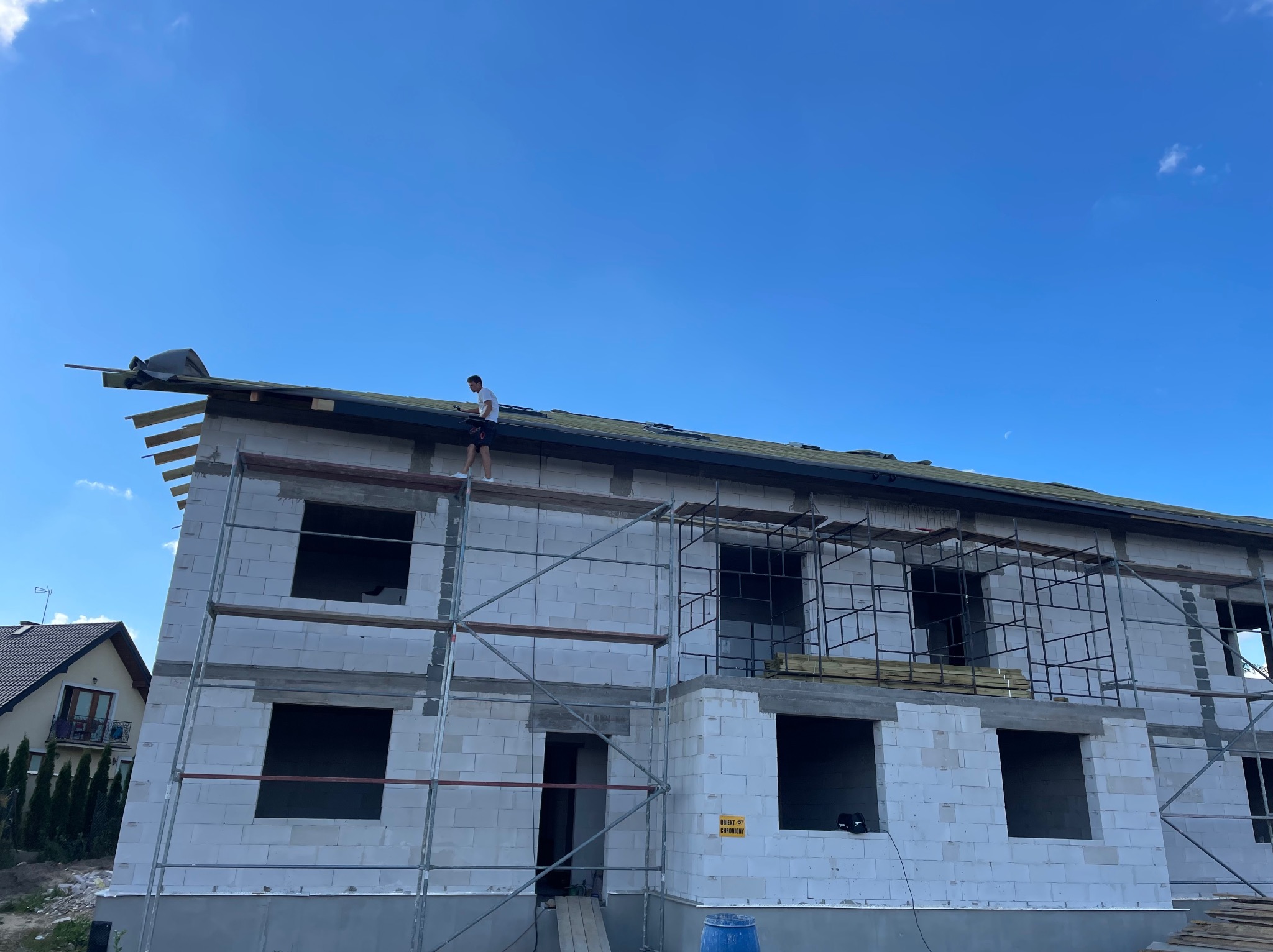 Budowa domu z białych bloczków: dekarz na dachu układa membranę dachową, widoczne rusztowania i niezabudowane okna.