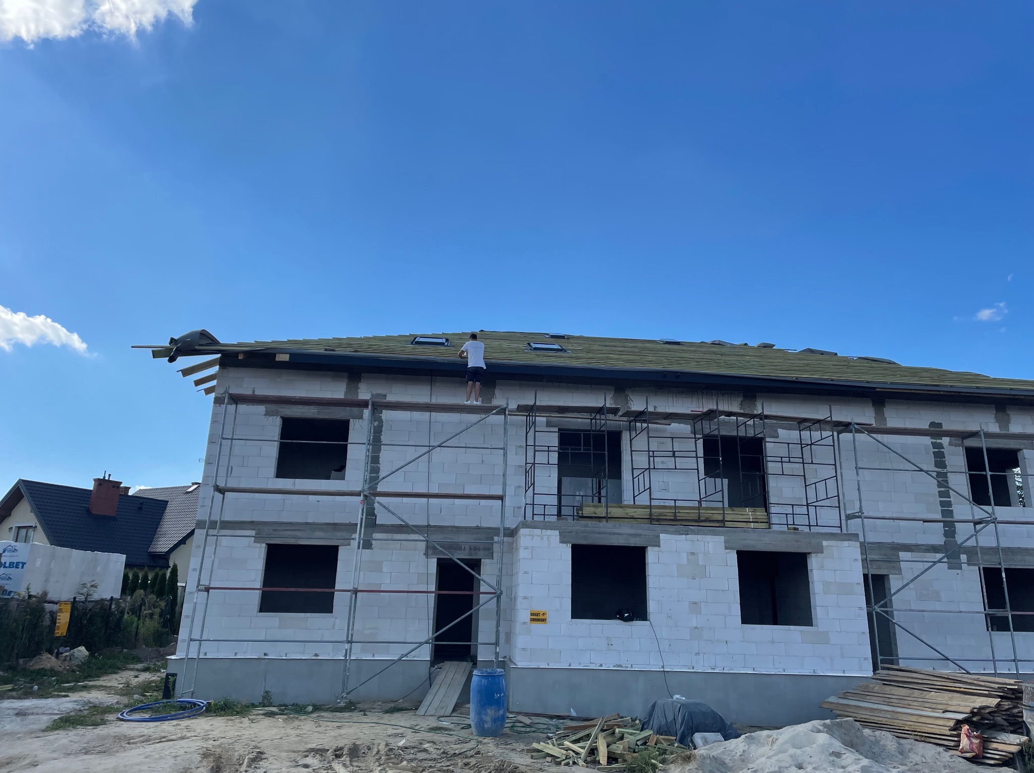 Wznoszony budynek z białego bloczka na etapie prac dekarskich, widoczne rusztowania i dekarz na dachu, błękitne niebo w tle.