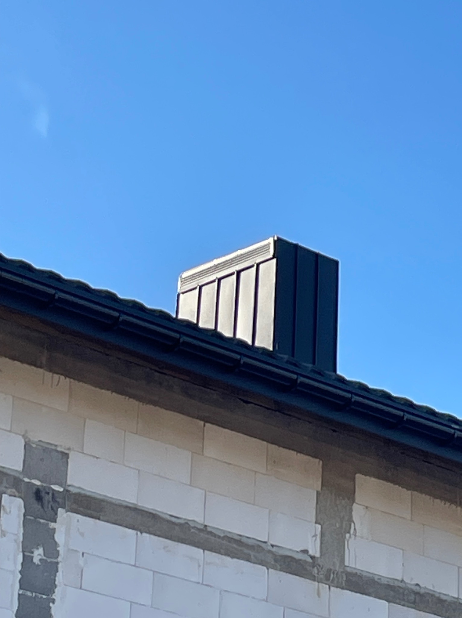 Niski kąt ukazuje czarny, metalowy komin na dachu budynku z białego bloczka, na tle błękitnego nieba.