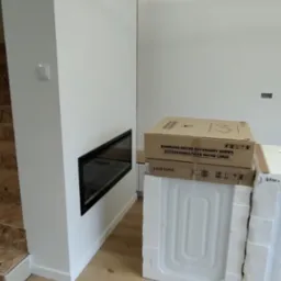 Nowoczesny salon w trakcie wykańczania: białe ściany, drewniana podłoga, wbudowany kominek i zapakowane urządzenia AGD Samsung czekające na montaż.