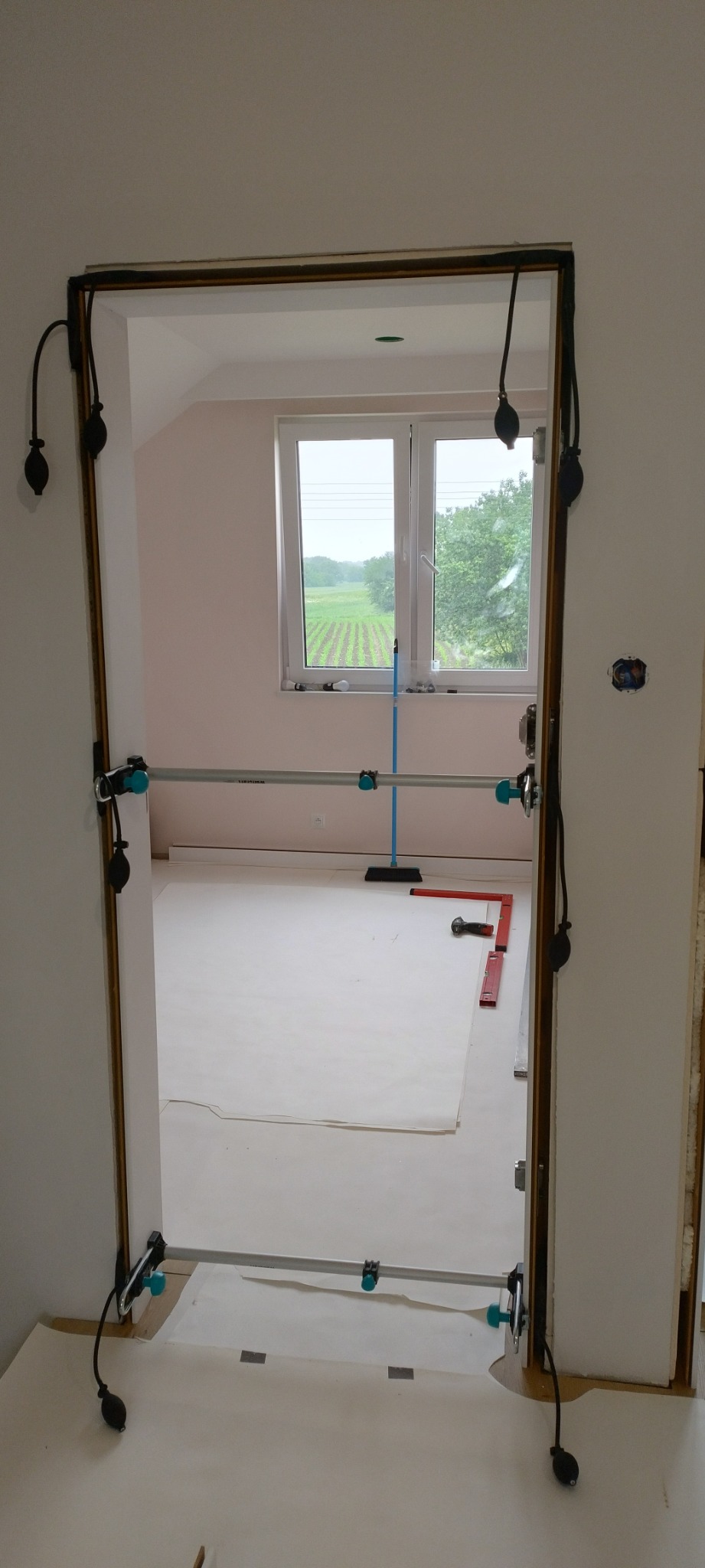 Montaż ościeżnicy regulowanej w przygotowanym otworze drzwiowym, widoczne narzędzia i poziomica, zabezpieczona podłoga i widok na pole za oknem.