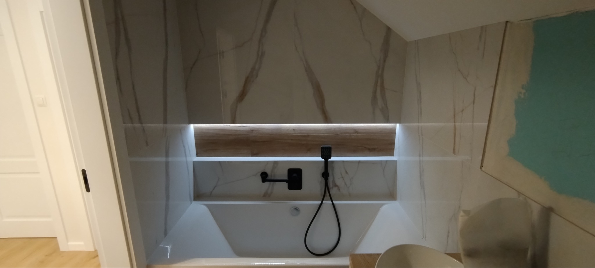 Nowoczesna łazienka z wanną w zabudowie, marmurowe płytki na ścianach, czarna armatura i podświetlana półka z drewnianym akcentem, fragment niedokończonej ściany po prawej stronie.