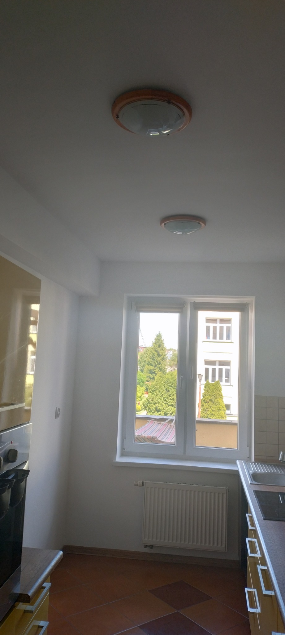 Jasna kuchnia z brązową terakotą na podłodze, widok na zieleń za oknem i dwa okrągłe plafony na suficie.