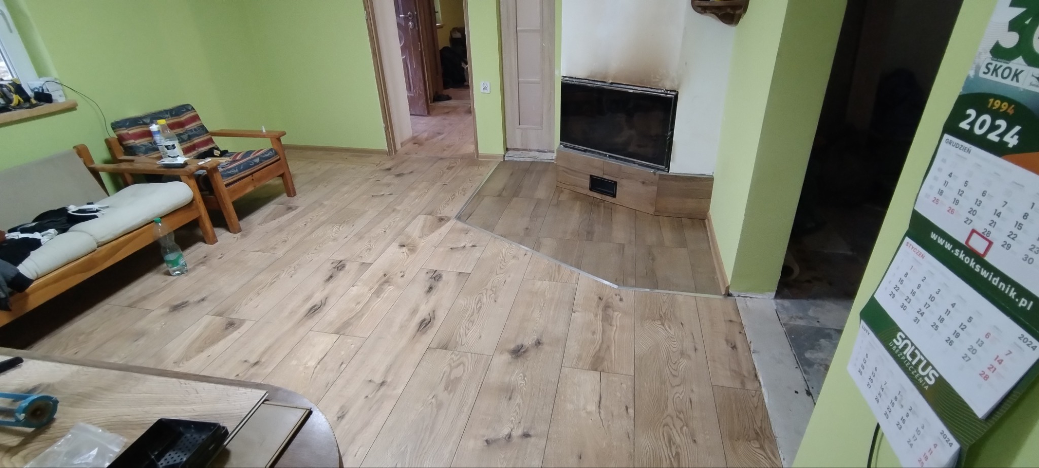 Ukończone układanie paneli podłogowych w pokoju z kominkiem, widoczne przejście do innego pomieszczenia i fragment umeblowania.