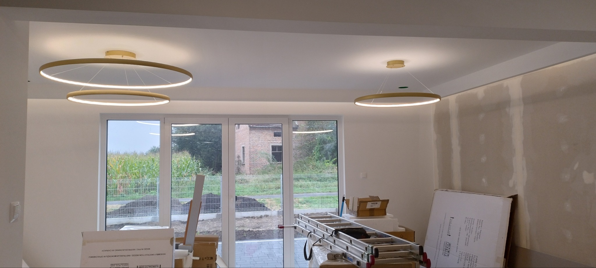 Wnętrze w trakcie wykańczania z trzema nowoczesnymi okrągłymi lampami LED, widoczne gładzie na ścianach, okno z widokiem na pole i drabiną opartą o ścianę.