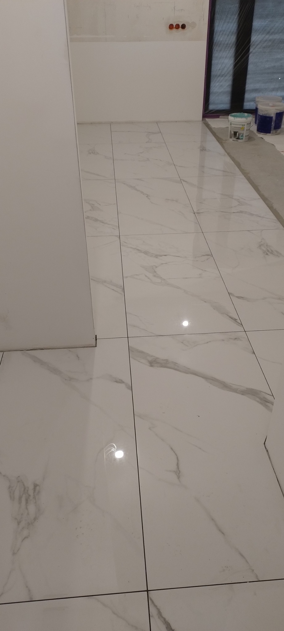 Białe płytki podłogowe imitujące marmur w trakcie układania, widoczne puszki instalacyjne w ścianie i wiaderka z farbą w tle.