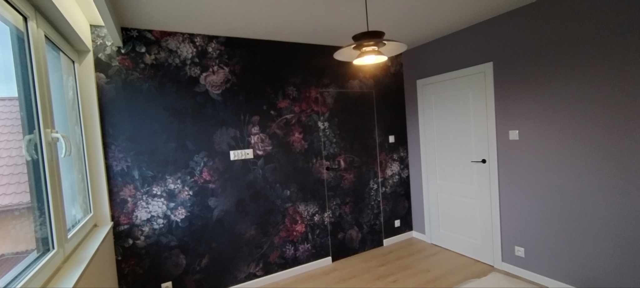 Pokój po remoncie z fototapetą w kwiaty na jednej ścianie, szara ściana, białe drzwi i okno, nowoczesna lampa wisząca.