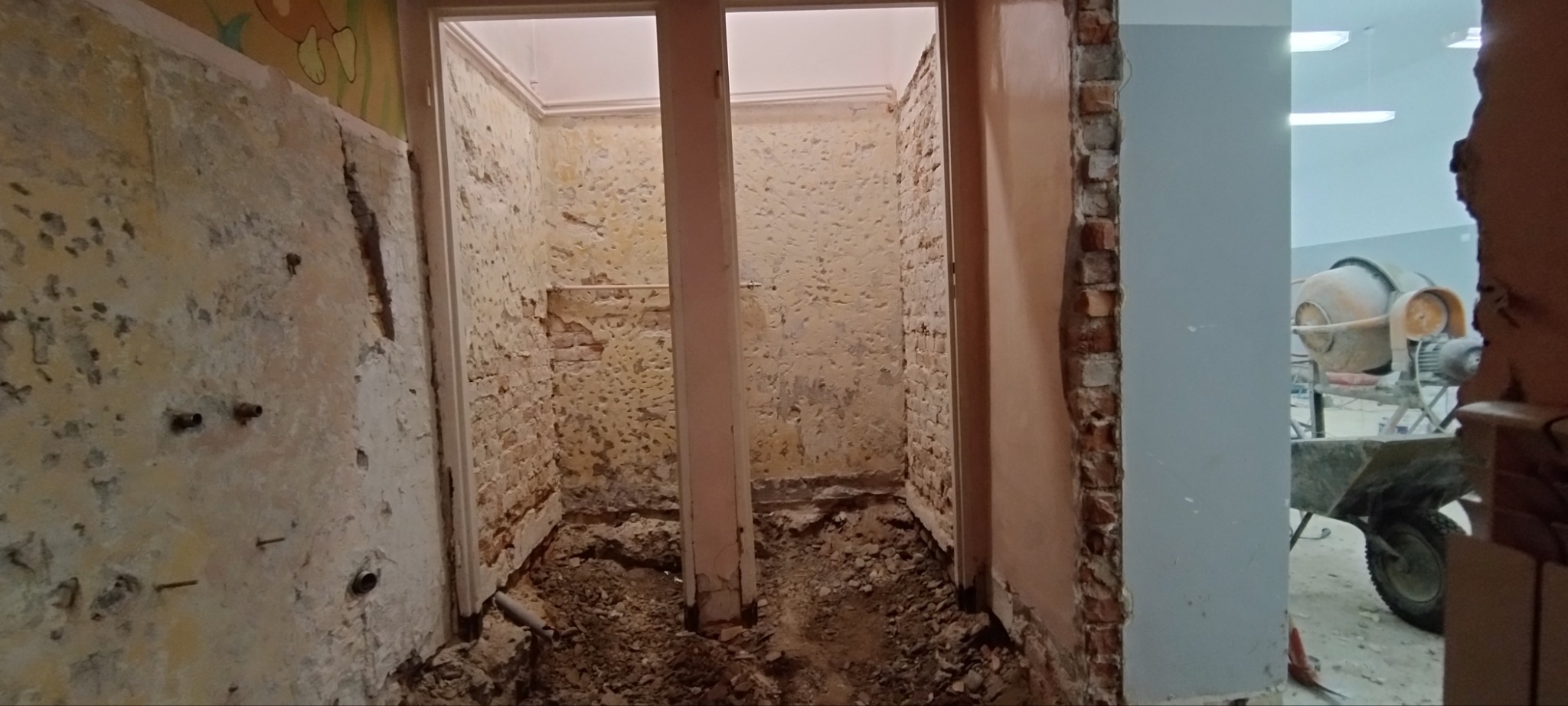Wnętrze w trakcie demontażu: odsłonięte cegły, resztki starej farby, gruz na podłodze, widoczny mikser do betonu w tle.