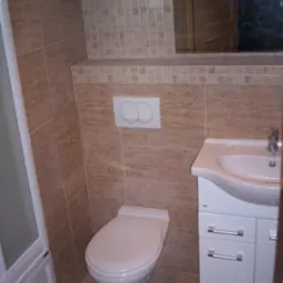 Wykończona łazienka z beżowymi płytkami, białą toaletą wiszącą i szafką pod umywalką z chromowanymi uchwytami.