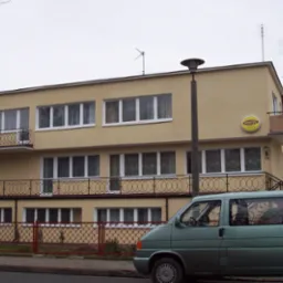 Dwupiętrowy budynek z beżową elewacją, balkonami z metalowymi balustradami i licznymi oknami, widziany z ulicy, z zaparkowanym zielonym vanem z reklamą firmy remontowej w tle.