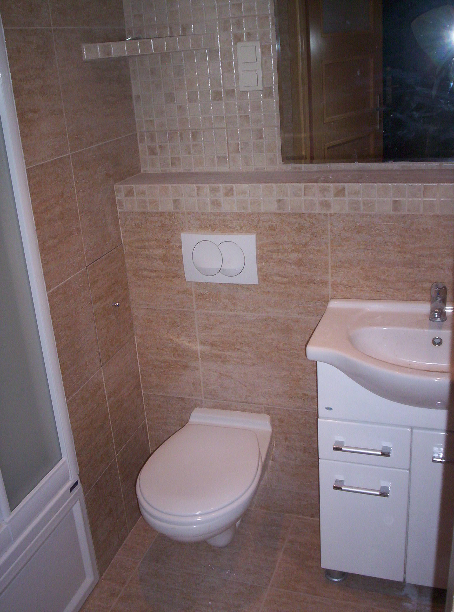 Wykończona łazienka z beżowymi płytkami, białą toaletą wiszącą i szafką pod umywalką z chromowanymi uchwytami.