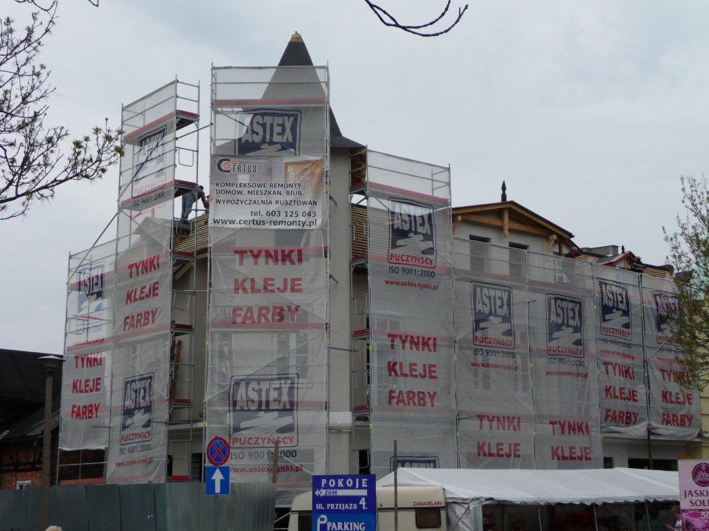 Budynek w trakcie renowacji, osłonięty rusztowaniami pokrytymi reklamami materiałów budowlanych, widoczny fragment pracującej osoby na rusztowaniu.