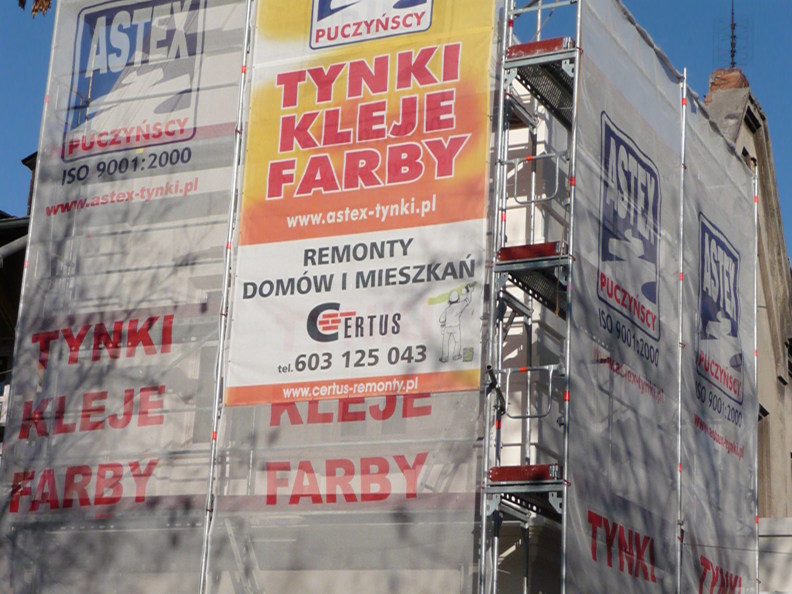 Elewacja budynku w trakcie remontu, osłonięta rusztowaniem z reklamami tynków, klejów i farb firmy Astex i Certus, widoczne drabiny rusztowania.