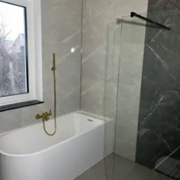 Nowoczesna łazienka z wanną, złotą armaturą i szarymi płytkami imitującymi marmur, oddzielona szklanym panelem z czarnym uchwytem.