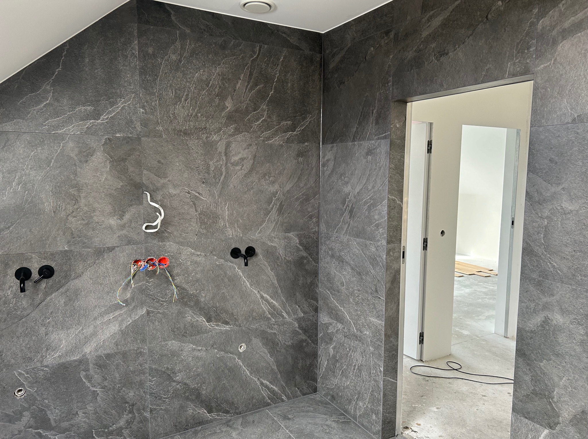 Łazienka w trakcie wykańczania: szare płytki imitujące kamień, czarna armatura, wystające przewody elektryczne, widok na sąsiednie pomieszczenie z panelami na podłodze.