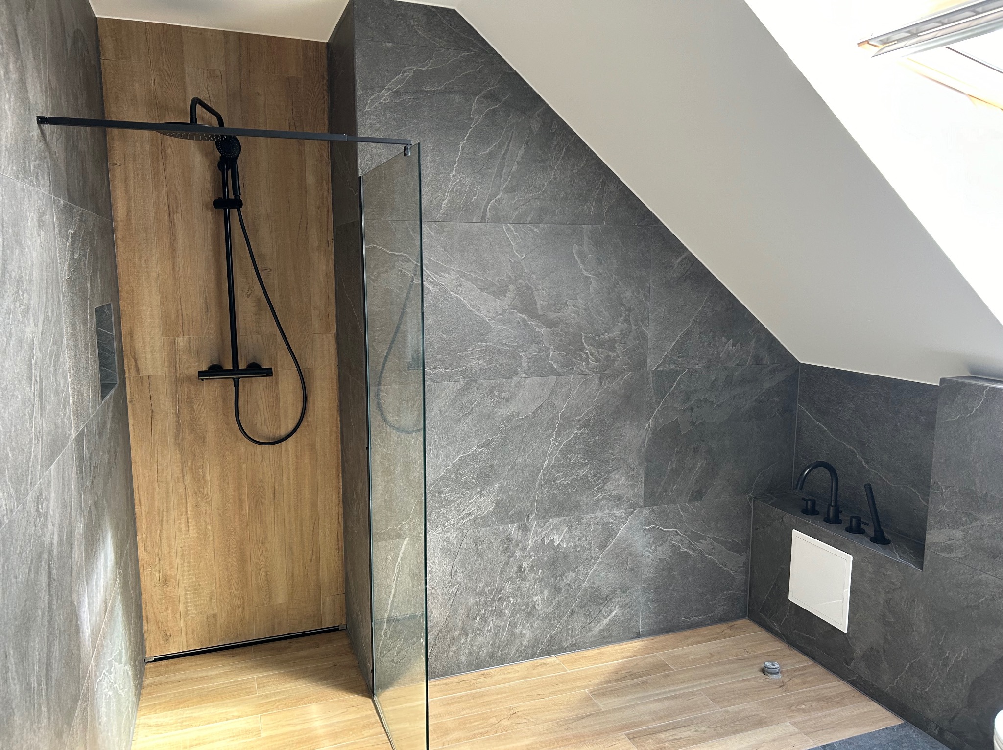 Nowoczesna kabina prysznicowa w łazience na poddaszu, z szarymi płytkami imitującymi kamień na ścianach i drewnopodobnymi płytkami na podłodze, czarna armatura i szklana ścianka kabiny.