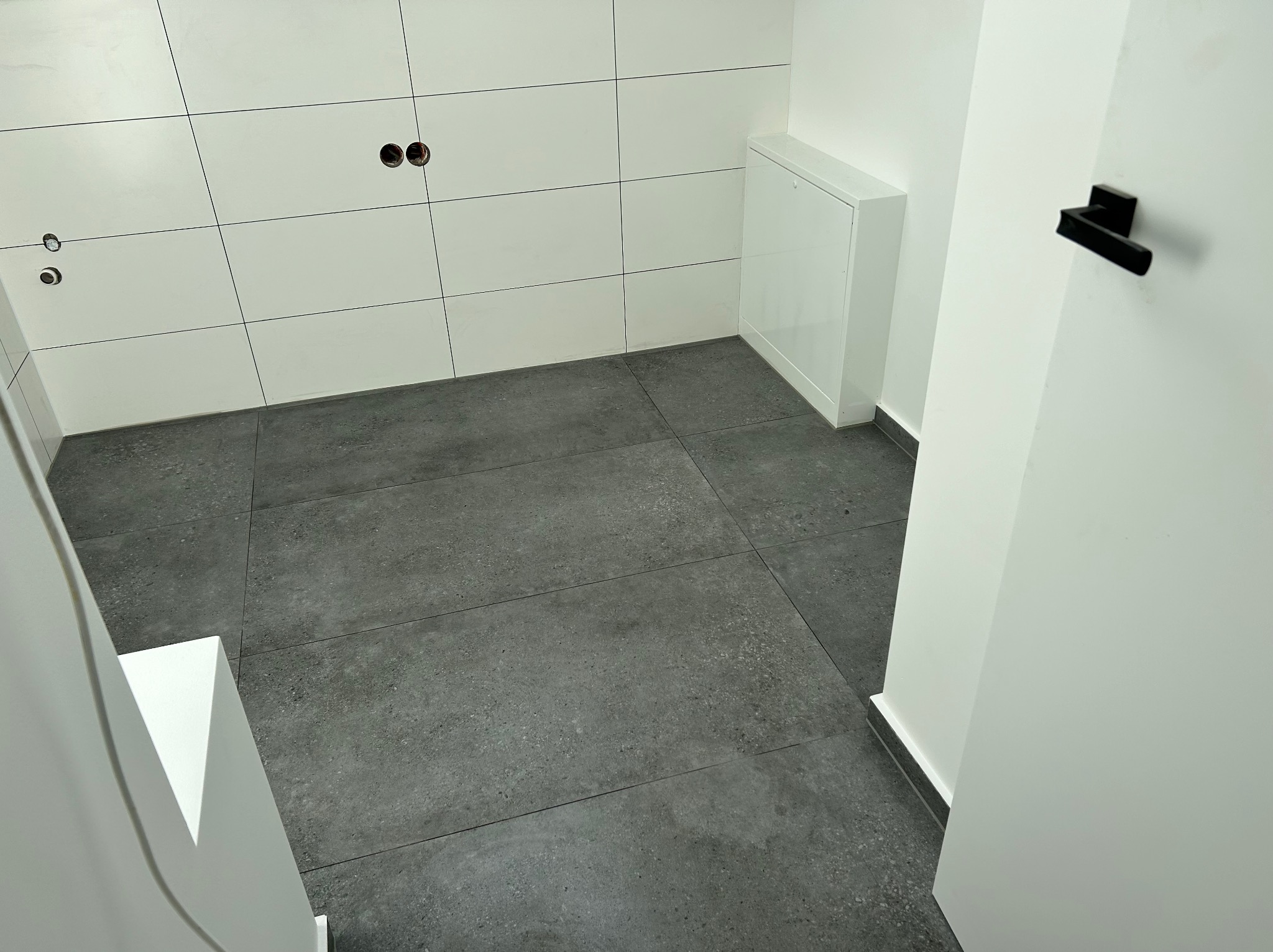 Nowoczesna łazienka w trakcie wykańczania: białe płytki ścienne z otworami na armaturę, szare płytki podłogowe, biała szafka i czarna klamka.